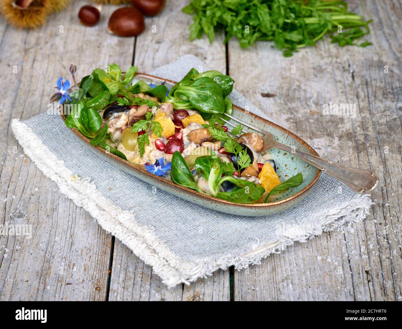 Insalata di castagne con uva, arancia, lattuga di agnello e melograno in una ciotola allungata su un panno, tavolo di legno non trattato con castagne, bucce e chervello Foto Stock