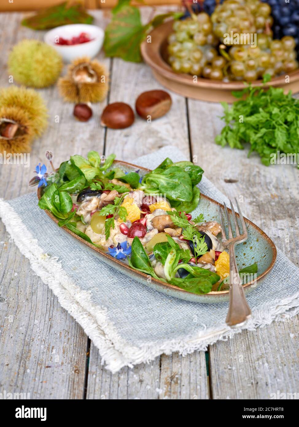 Insalata di castagne con uva, arancia, lattuga di agnello e melograno in una ciotola allungata su un panno, tavolo di legno non trattato con castagne, bucce e chervello Foto Stock