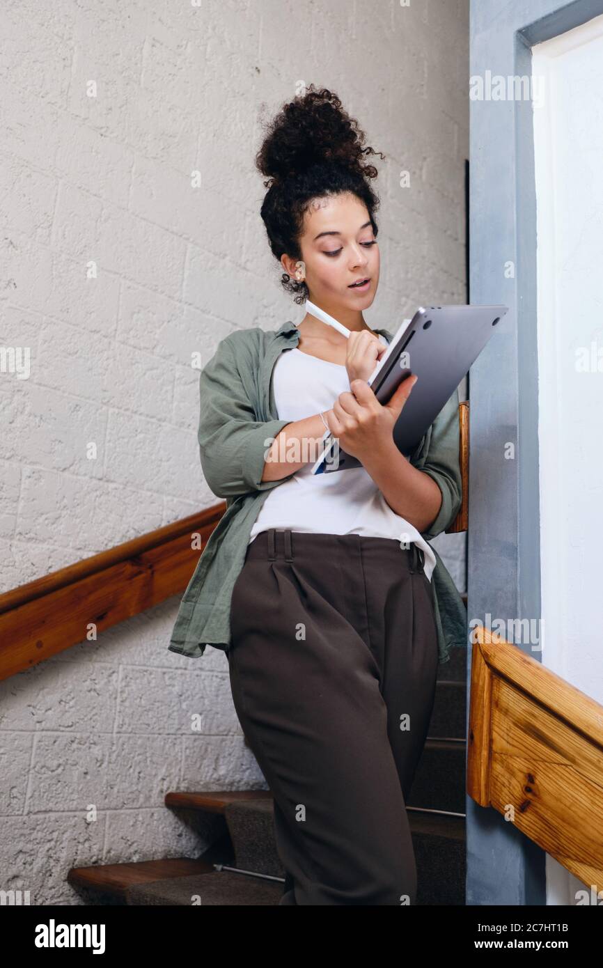 Giovane ragazza studentesca attraente con capelli ricci scuri in camicia kaki e pantaloni in piedi sulle scale in università tenendo il laptop in mani pensierose Foto Stock