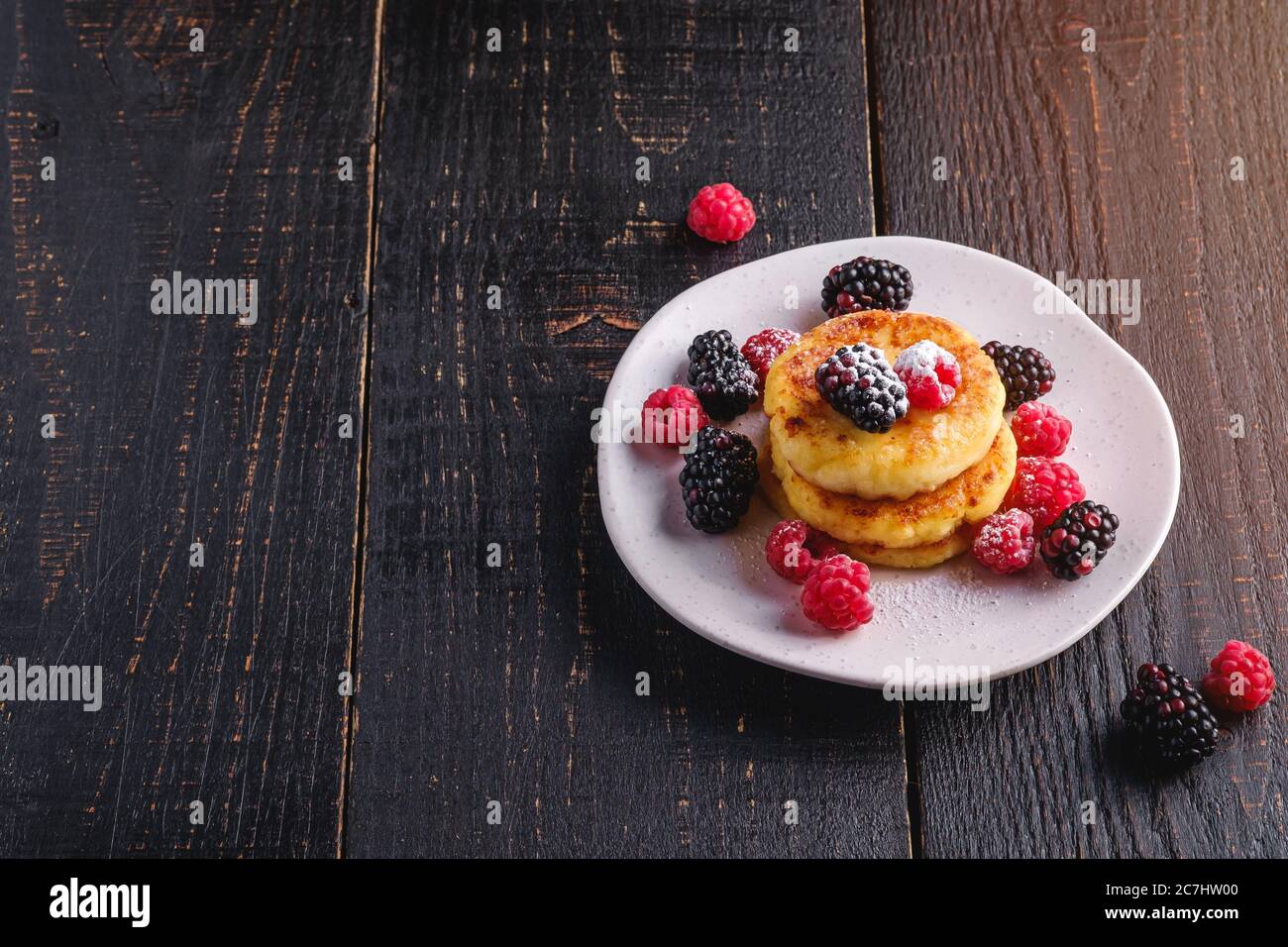 Frittelle di formaggio e zucchero in polvere, frittelle di cagliata dessert con lampone e frutti di bosco in piatto su sfondo nero scuro di legno Foto Stock