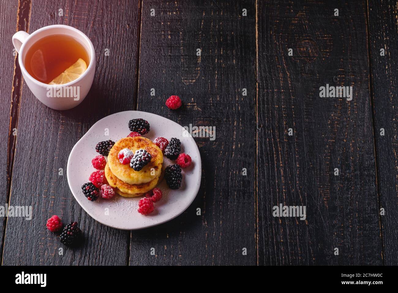 Frittelle di formaggio cottage, frittelle di cagliata dessert con lampone e frutti di bosco in piatto vicino alla tazza di tè caldo con fetta di limone Foto Stock
