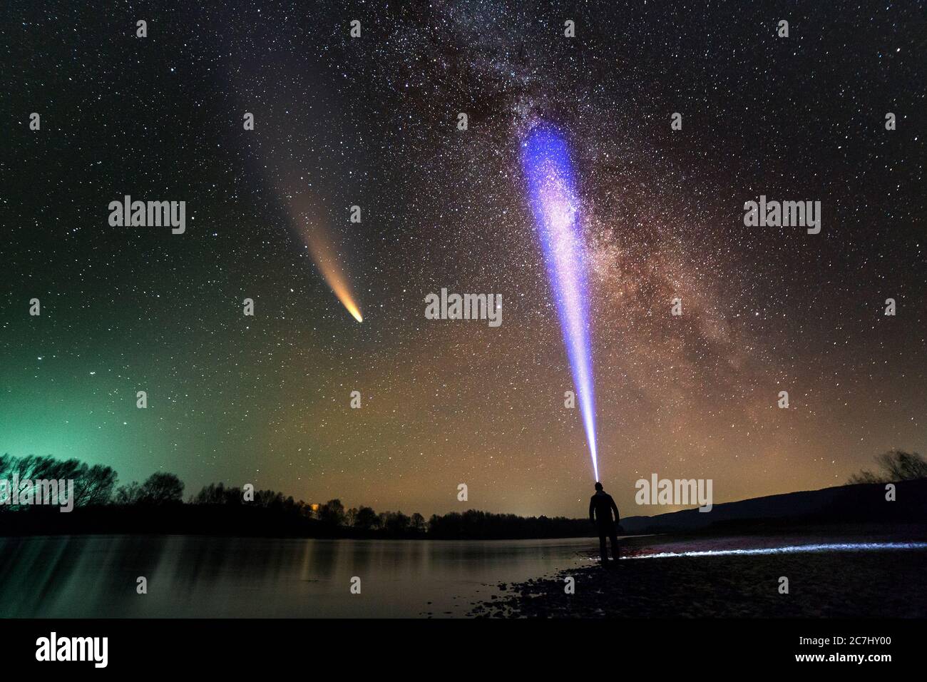 Silhouette di un uomo con torcia da testa in piedi sulla riva del fiume che illumina un fascio di luce sulla galassia di Milkyway e la cometa di Neoswise con coda chiara nel buio Foto Stock