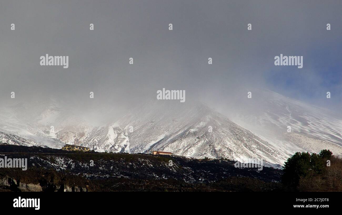 Etna, Parco Nazionale dell'Etna, Parco dell'Etna –´Etna, pendio di lava nera in primo piano, Rifugio Sapienza al centro, nuvole grigie sopra, oltre l'Etna innevata Foto Stock