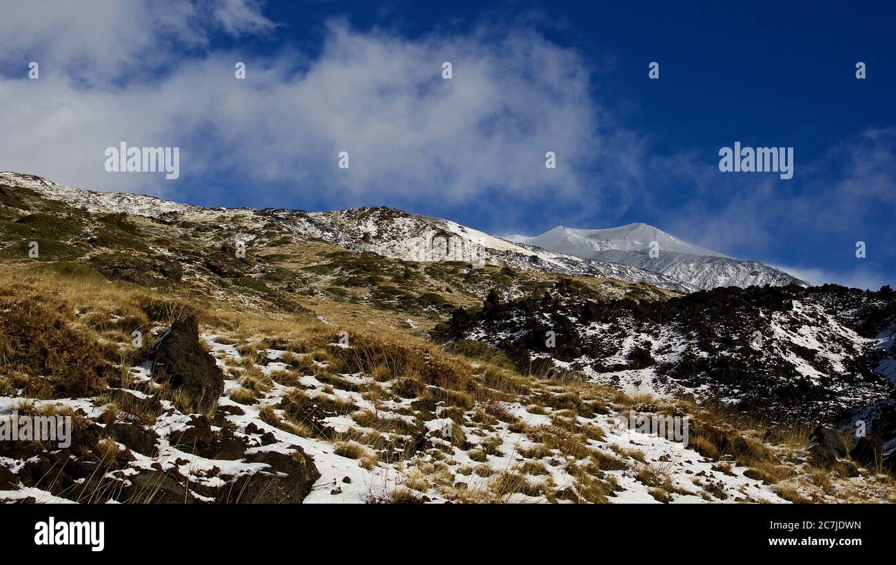 Etna, Parco Nazionale dell'Etna, Parco dell'Etna –´Etna, paesaggio lavico giallo in primo piano, festa lavica nera al centro, cime innevate dell'Etna sullo sfondo, cielo più calvo con nuvole bianche Foto Stock