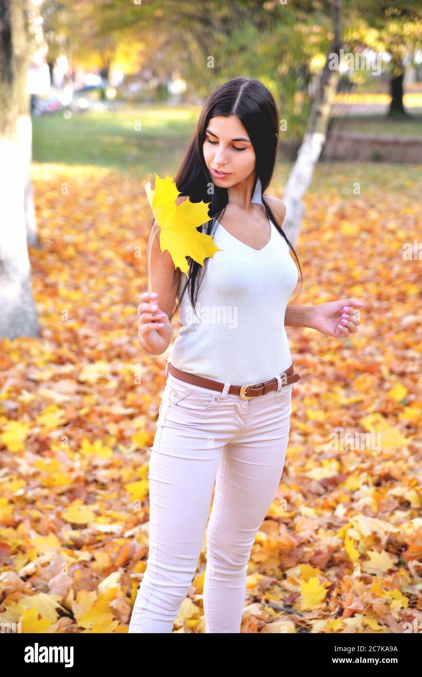 Bella ragazza in parco autunno tenendo una foglia d'acero in mano, sorridendo sul suo volto, in jeans bianchi e una blusa, su uno sfondo di alberi e voi Foto Stock