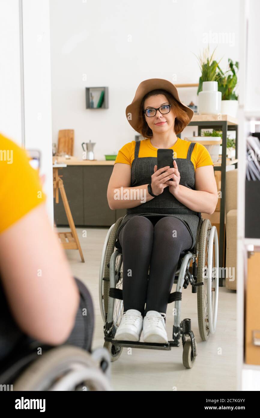Giovane donna abbastanza disabile in casualwear e occhiali facendo selfie in sedia a rotelle davanti allo specchio mentre provando su nuovo cappello a casa Foto Stock