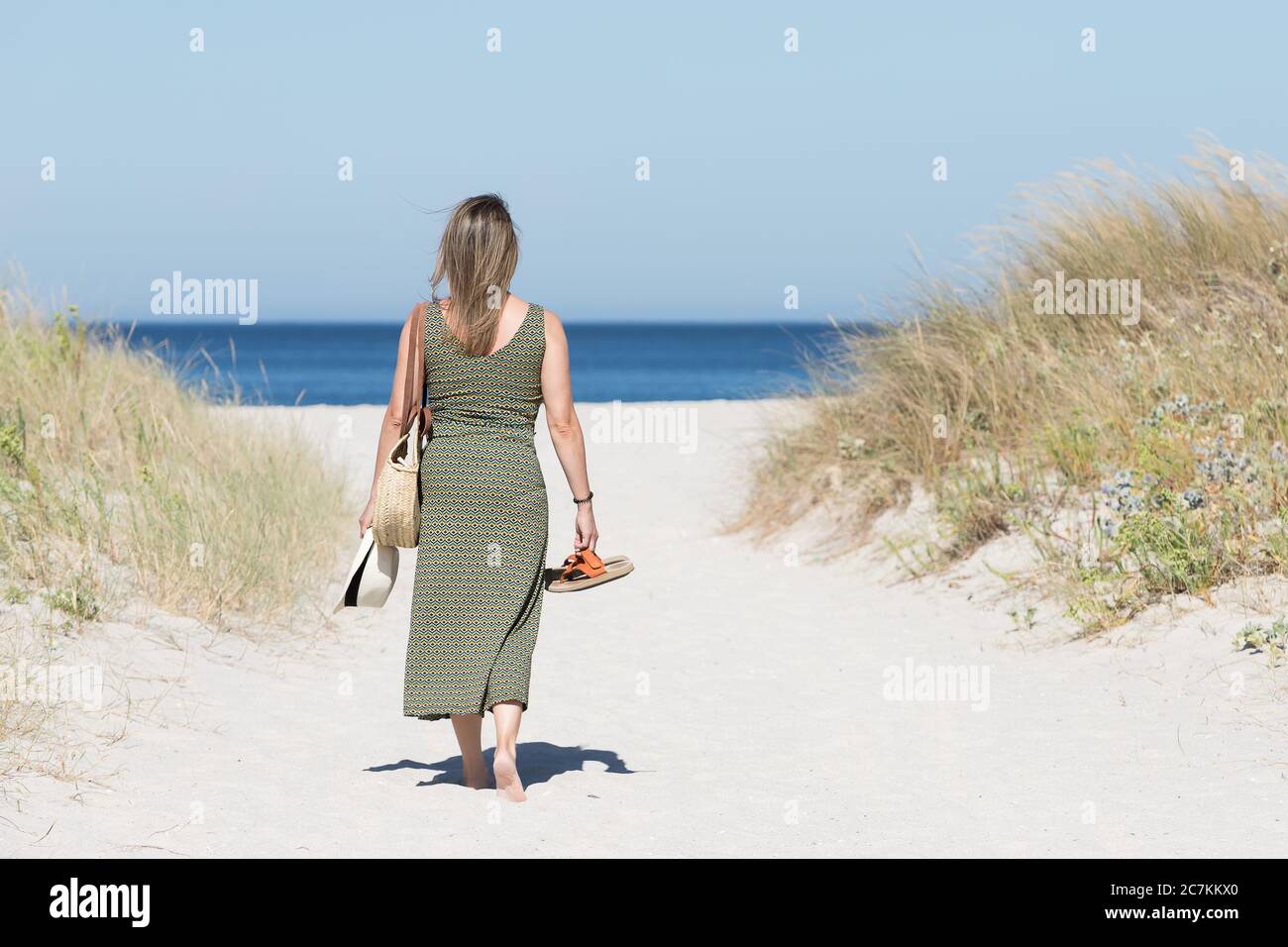 Donna bionda di mezza età in un abito a piedi nudi lungo un percorso sabbioso verso il mare con i suoi sandali in una mano e un cappello nell'altra. Foto Stock