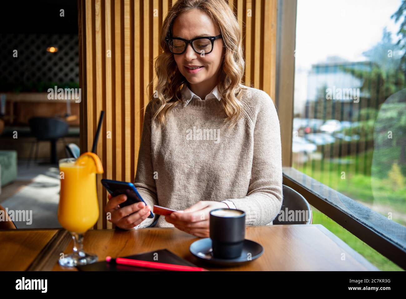 Elegante donna bionda seduta al caffè che tiene il telefono cellulare e la carta di credito che si iscrono sul sito web. Donna d'affari che paga con carta di credito mentre acquista Foto Stock
