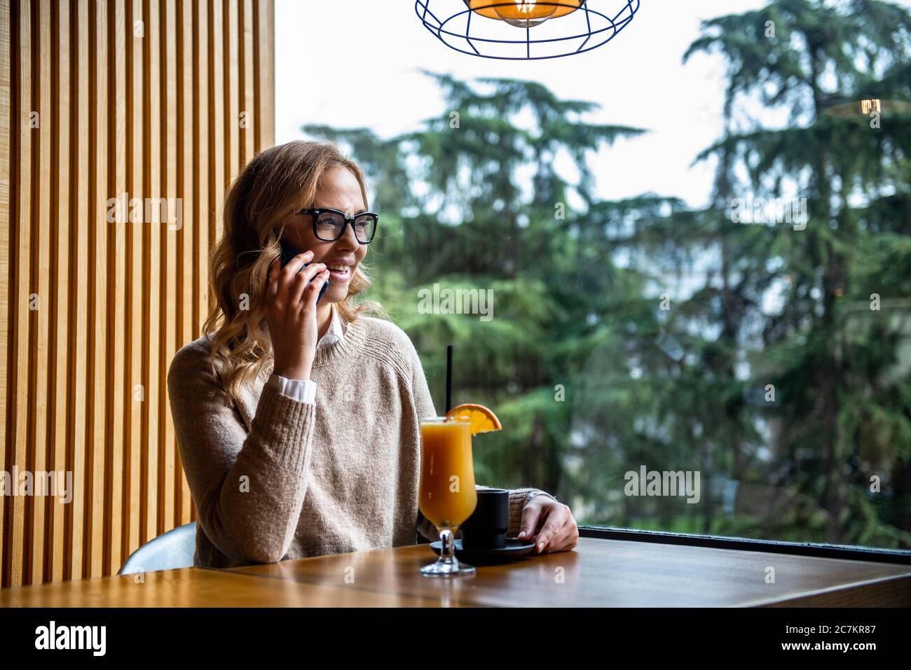 Giovane donna affascinante chiamare con il telefono cellulare mentre è seduto da solo in una caffetteria durante il tempo libero, attraente femmina con sorriso carino avere parlando Foto Stock