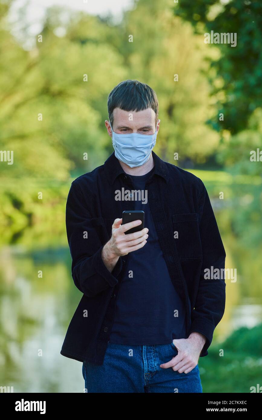 Uomo con maschera e cellulare in un parco cittadino, crisi di Corona, Ratisbona, Baviera, Germania Foto Stock