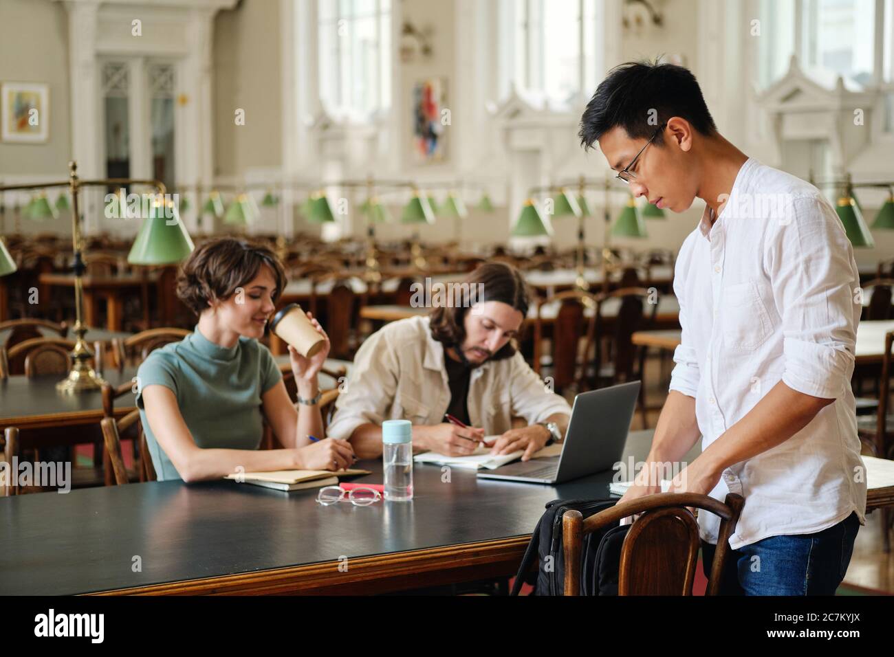 Giovane studente asiatico in occhiali con zaino che viene a studiare con gli amici in biblioteca di università Foto Stock