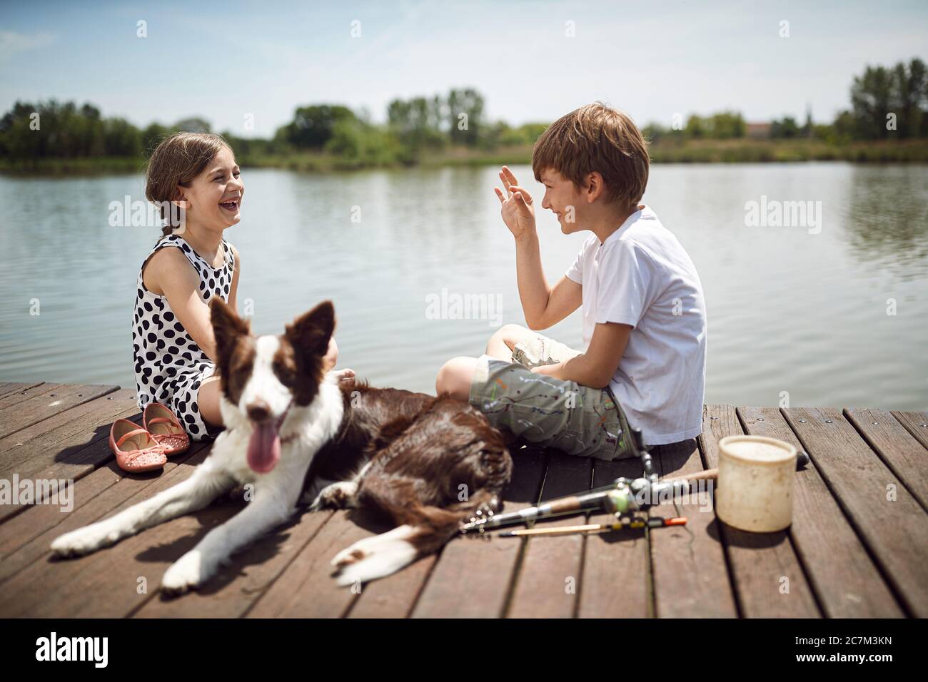 Felice infanzia in nature.smiling ragazzo e ragazza con cane seduto insieme su legno vicino stagno e parlare. Foto Stock