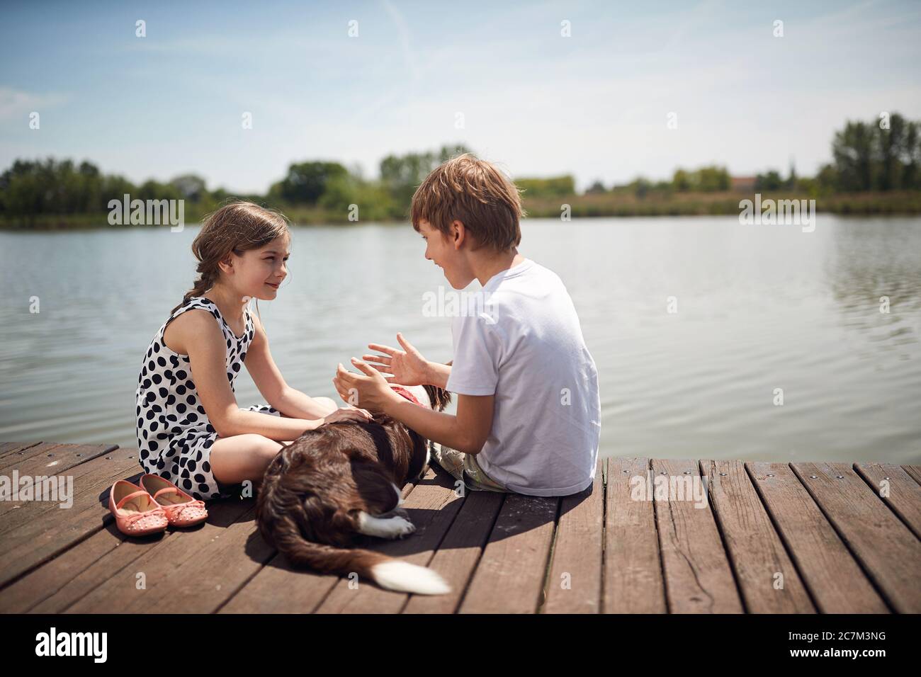 Infanzia felice in nature.Little bambini con cane seduta insieme su legno vicino stagno e parlare. Foto Stock