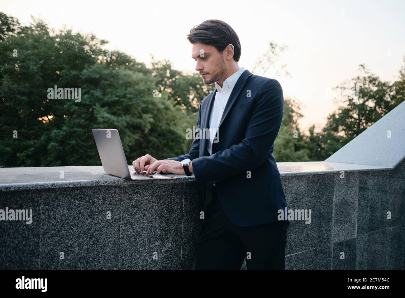 Giovane e attraente uomo in tuta classica in piedi per strada e lavorando con cura su un computer portatile Foto Stock