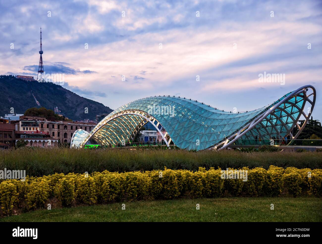 Ponte della Pace sullo sfondo del Monte Mtatsminda e la torre della TV. Paesaggio urbano di Tbilisi in serata al tramonto Foto Stock