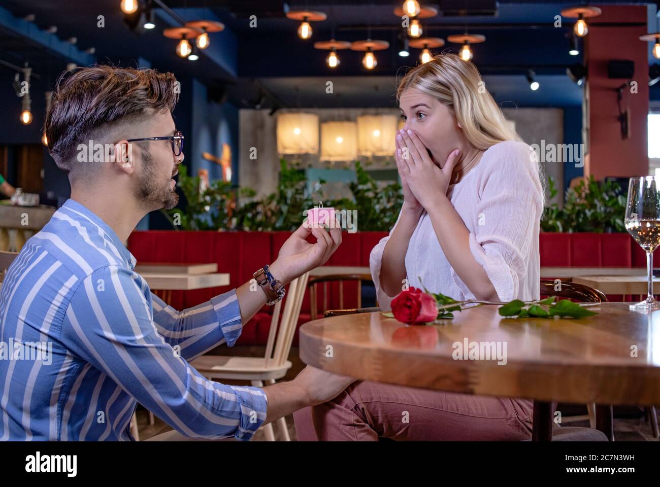 Giovane coppia romantica in un bel ristorante di fantasia. Il ragazzo sorprende la sua ragazza con una rosa rossa e le propone con un anello di fidanzamento. Foto Stock