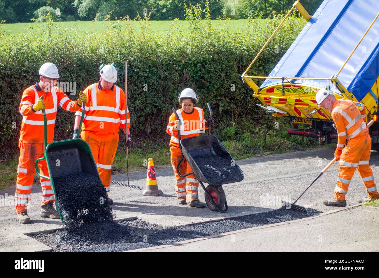 Operai e donne che fanno riparazioni stradali riparando la superficie stradale irregolare danneggiata e buchi di pentola con asfalto in una strada di paese di Cheshire Inghilterra UK Foto Stock