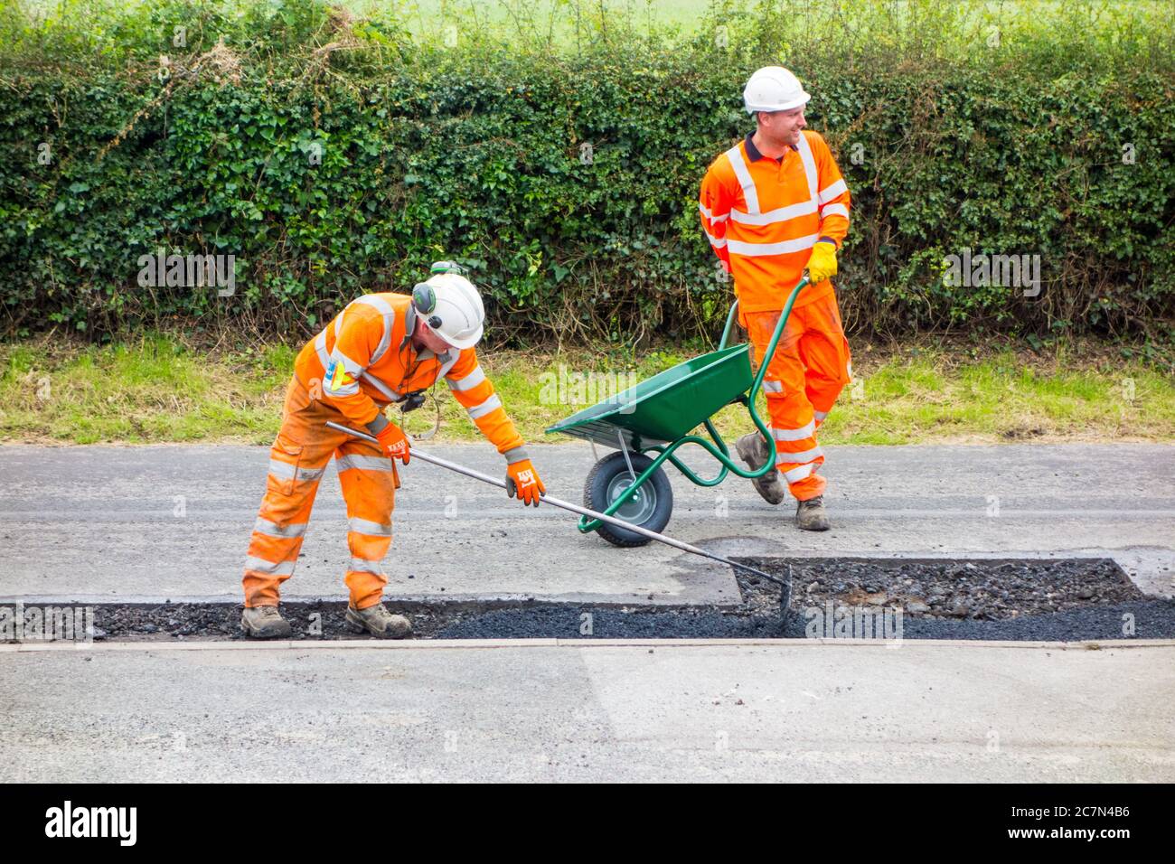 Operai e donne che fanno riparazioni stradali riparando la superficie stradale irregolare danneggiata e buchi di pentola con asfalto in una strada di paese di Cheshire Inghilterra UK Foto Stock