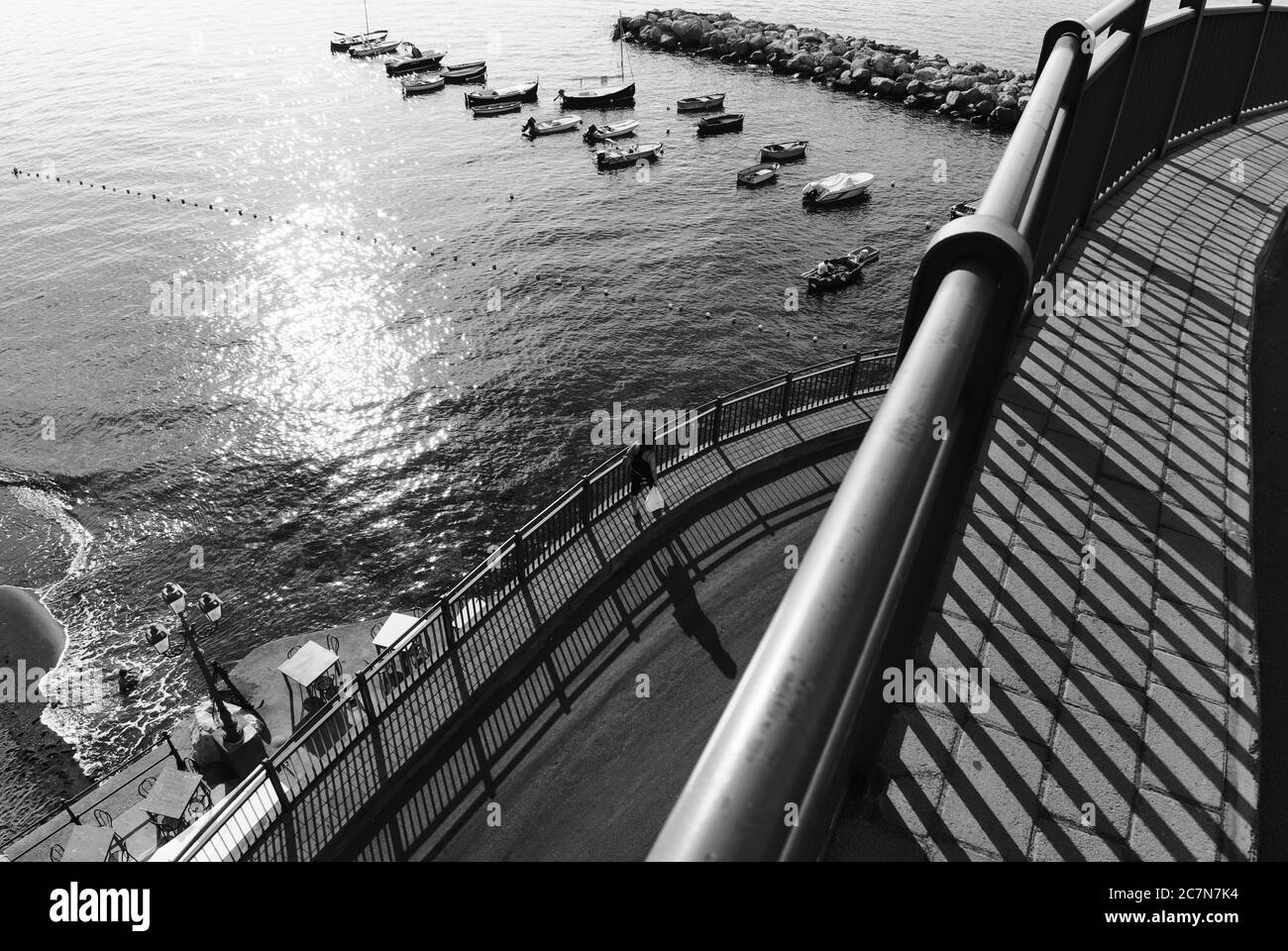 Il sole in bianco e nero riflette sul mare, i dettagli dell'architettura, le ringhiere getta lunghe ombre sul marciapiede dove una donna cammina lungo Amalfi C. Foto Stock
