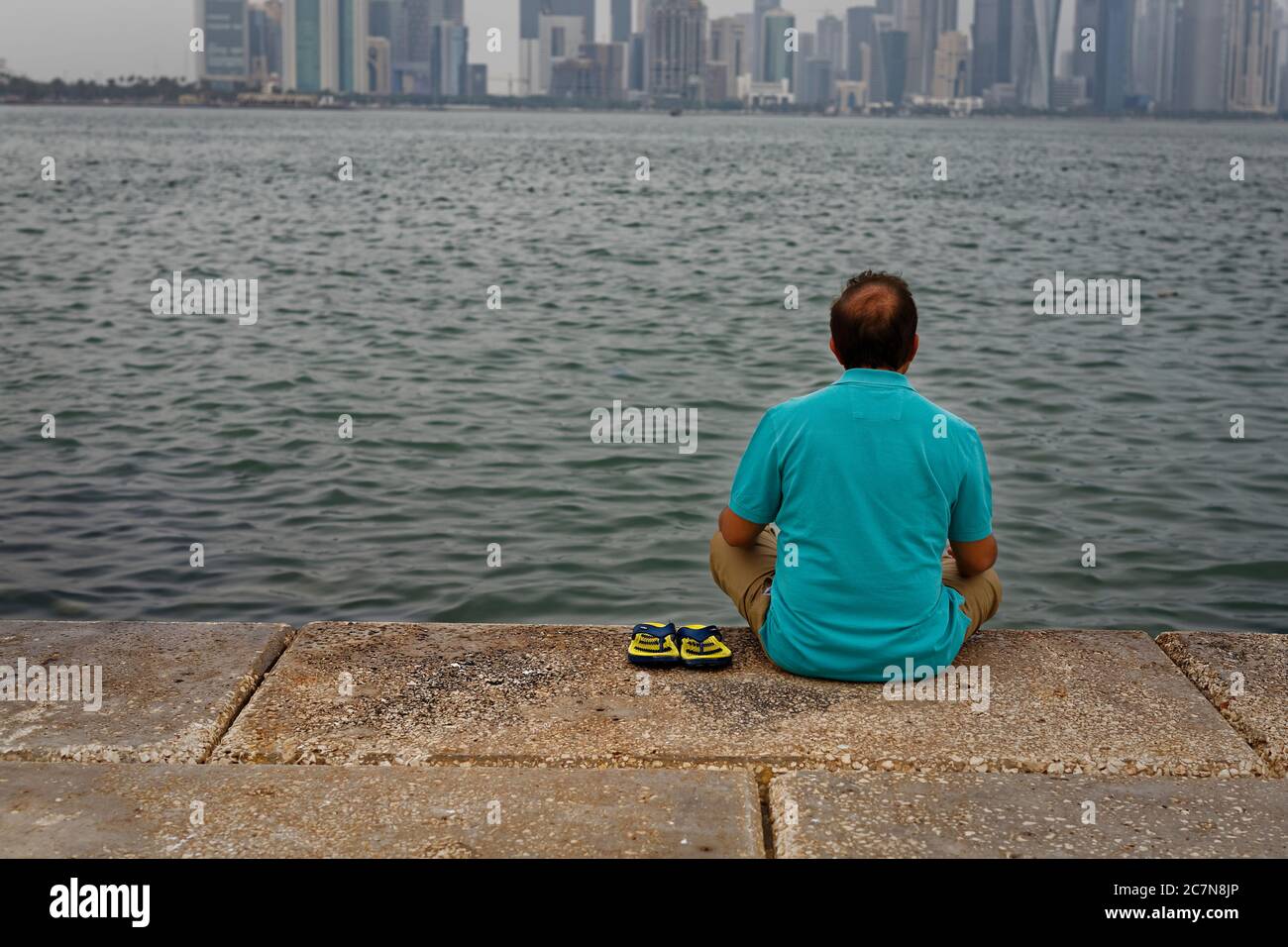 Un uomo seduto sulla nicchia di Doha cor e guardando al golfo arabo e grattacieli fotografati da dietro in luce del giorno Foto Stock