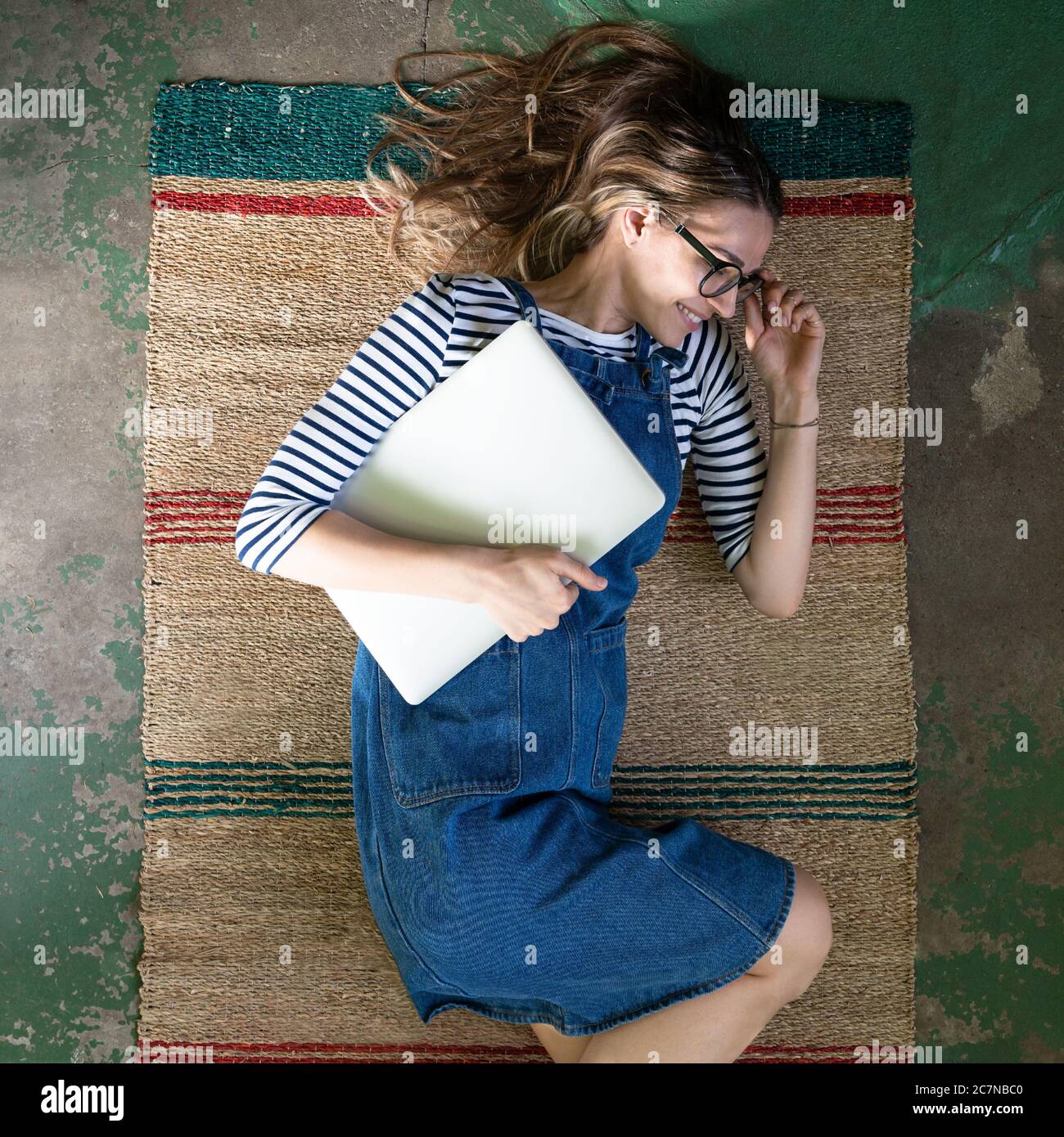 Vista dall'alto della donna carina in occhiali indossare tute in jeans blu, giacente su tappeto di canna sul pavimento in cemento, tenendo il laptop e riposandosi dopo il lavoro. Foto Stock