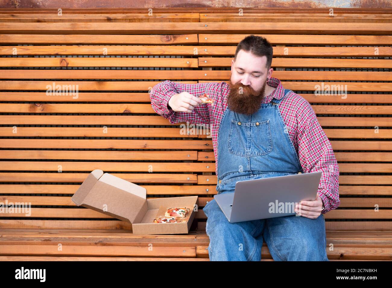 Uomo hipster indolente e indolente in tute in jeans blu, maglietta controllata che lavora su un computer portatile e che mangia pizza da asporto, seduto su una panca. Fast food Foto Stock