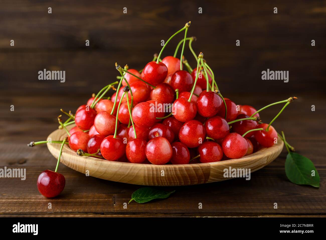 Ciliegie rosse dolci in un piatto di legno e foglie verdi su un tavolo di legno marrone. Messa a fuoco selettiva. Foto Stock
