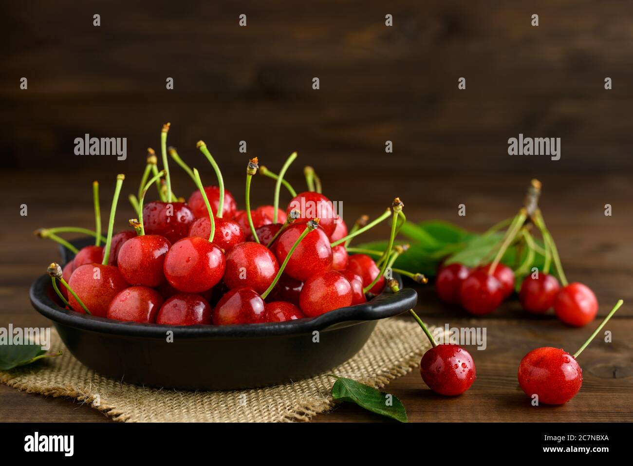 Ciliegie rosse dolci fresche in una ciotola nera e foglie verdi su un tavolo di legno marrone scuro. Messa a fuoco selettiva. Foto Stock
