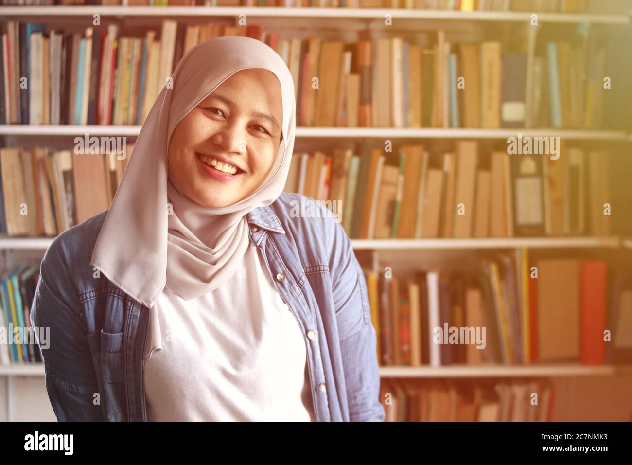 Ritratto di allegra libricana musulmana asiatica che indossa hijab, guardando la macchina fotografica e sorridendo, donna in piedi contro i libri in biblioteca Foto Stock