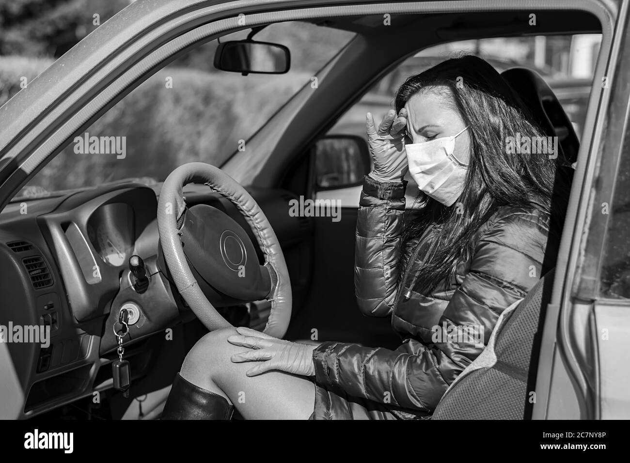 una ragazza in un'auto in una maschera medica anti virus con scarsa salute tiene la testa con la mano Foto Stock