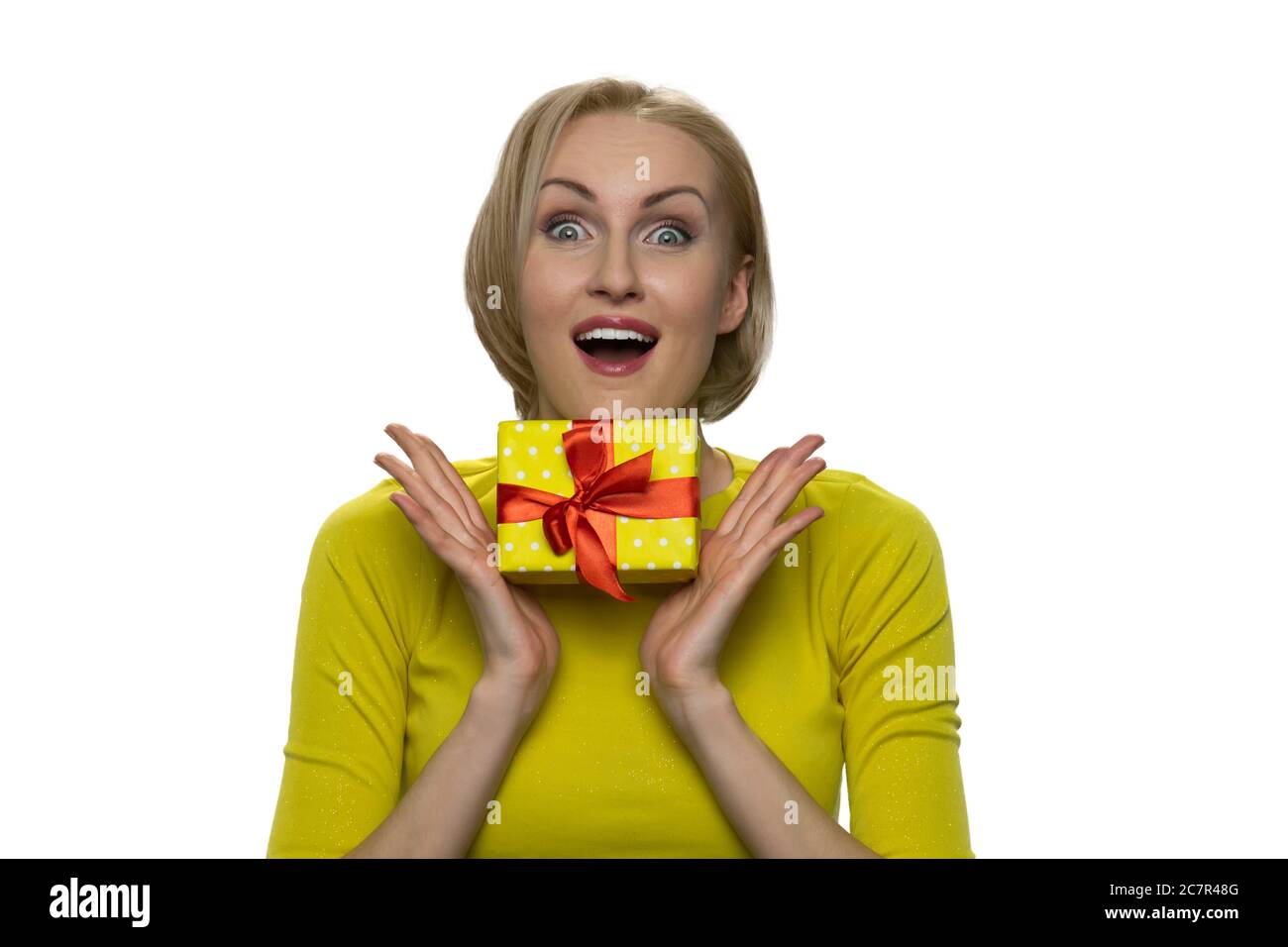 Donna bionda in dolcevita giallo preme il regalo al petto e guardando la macchina fotografica con gli occhi ampiamente aperti. Foto Stock