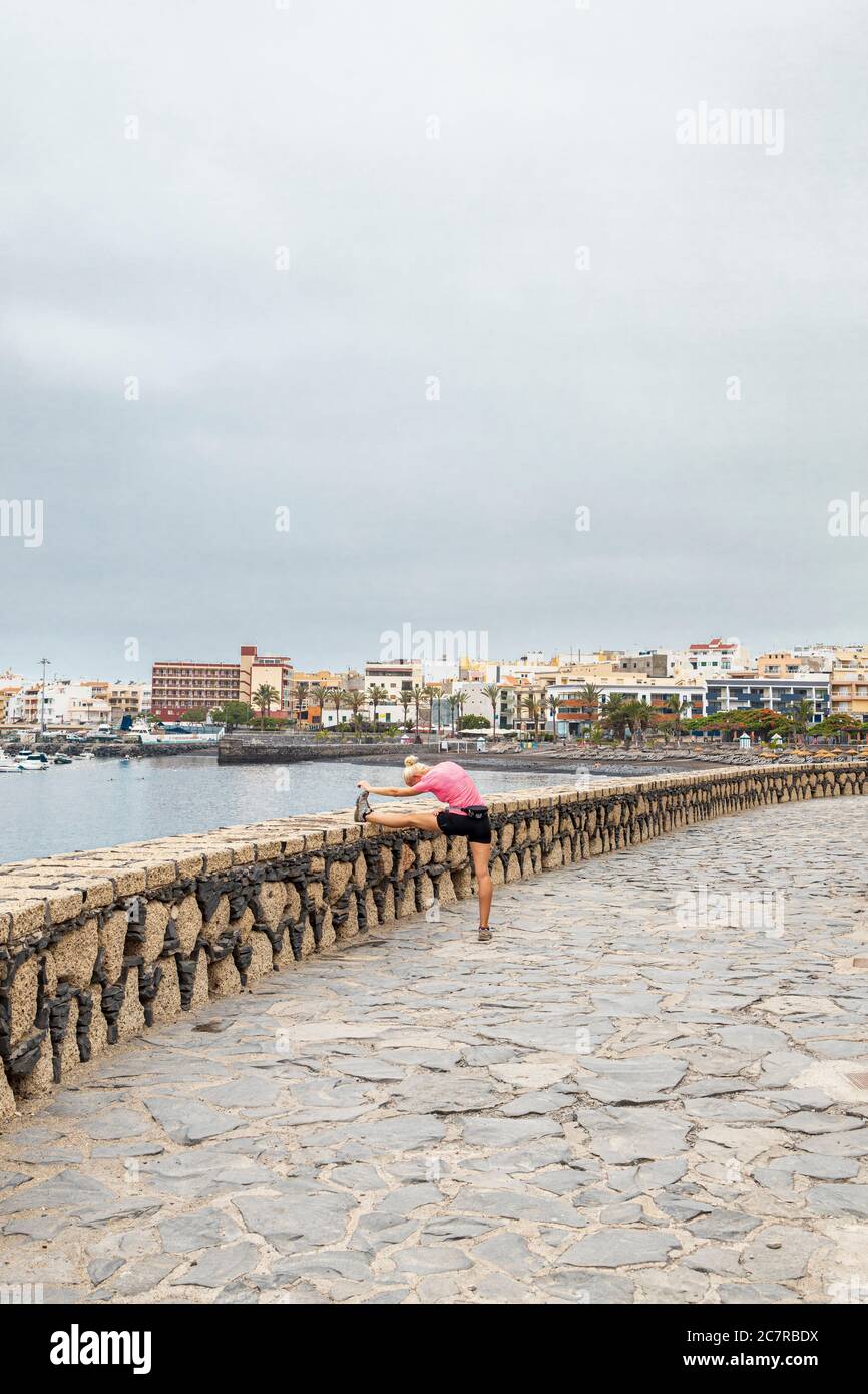Donna che stende i muscoli delle gambe sul muro lungo la passeggiata a Playa San Juan, Tenerife, Isole Canarie, Spagna Foto Stock