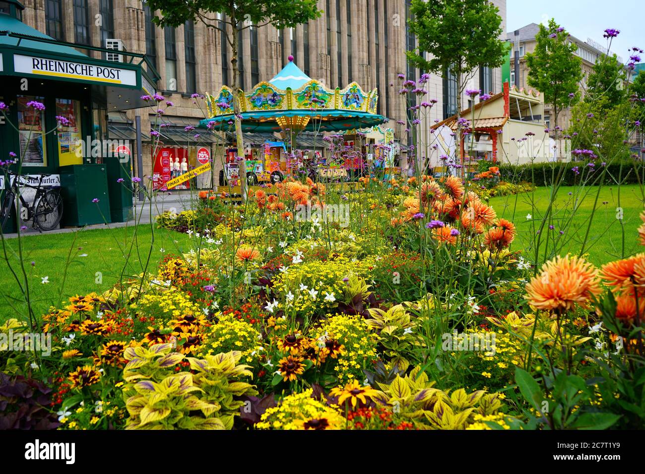 Vista attraverso i colorati fiori estivi: Corneliusplatz nel centro di Düsseldorf con edicola e una carosello nostalgico. Foto Stock