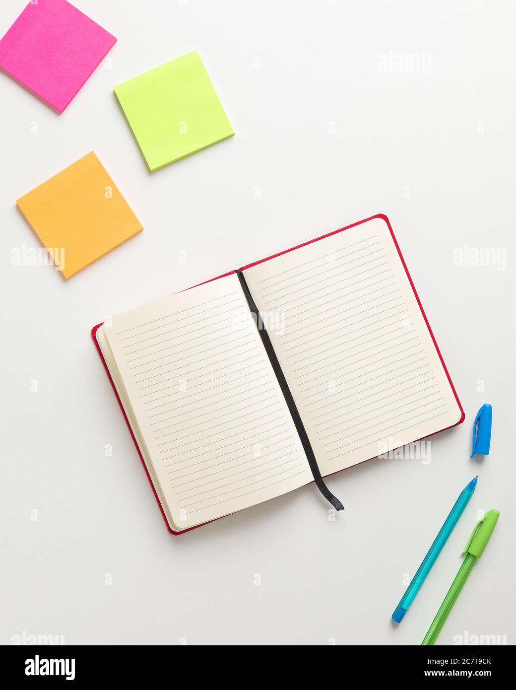 Vista dall'alto di un notebook rosso vuoto al centro, promemoria nell'angolo superiore e penna blu e verde nell'angolo inferiore, con sfondo bianco Foto Stock