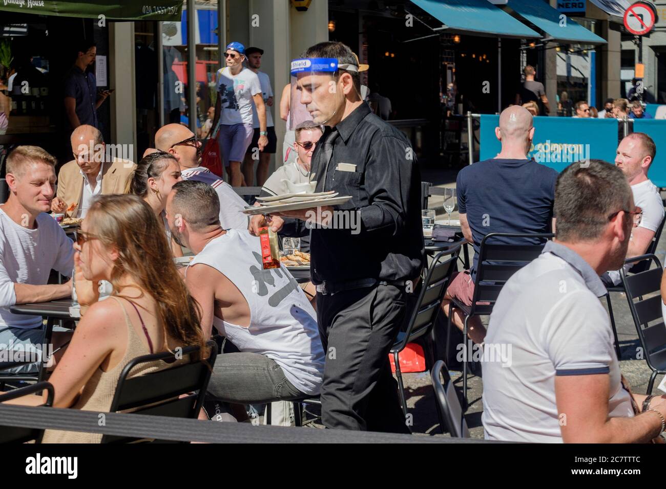 18 luglio 2020. Le strade nell'area di Soho nel centro di Londra sono state pedonali per consentire ai bar e ai ristoranti di allestire aree salotto all'aperto per mantenere le normative di allontanamento sociale tra i clienti, poiché le misure di allentamento Covid-19 sono state attuate dal governo: Un cameriere che indossa una protezione protettiva per il viso serve i clienti in un'area salotto all'aperto in Old Compton Street, Soho, Londra, Regno Unito. Foto Stock