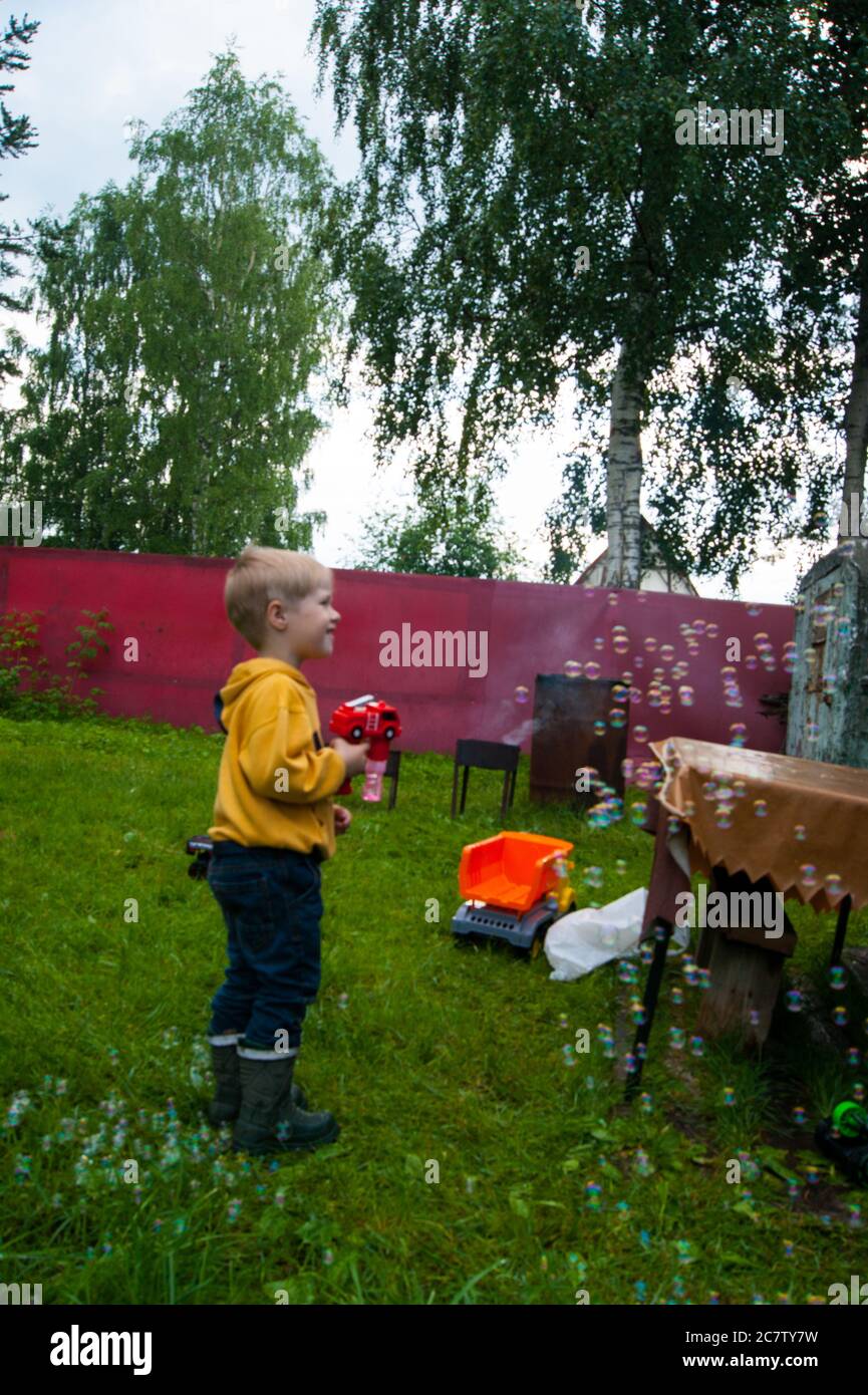 Un bambino e una pistola sotto forma di un camion fuoco rosso, che spara bolle di sapone in estate sul prato di campagna. Riposo, vacanza, animazione, bambini Foto Stock