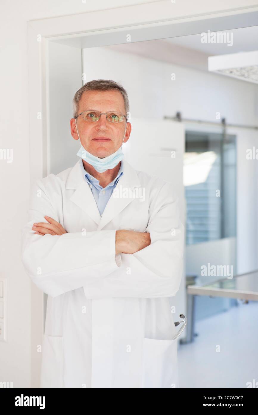 Medico sorridente maturo con maschera facciale in piedi con sicurezza di fronte alla sala operatoria - mettere a fuoco sul viso Foto Stock