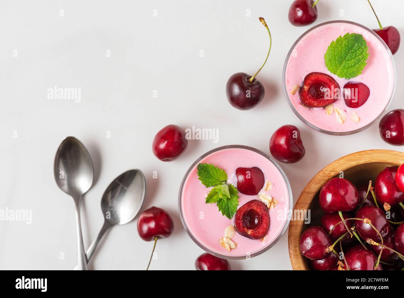 Colazione con dieta sana. Yogurt alla ciliegia in bicchieri con frutti di bosco freschi, avena, menta e cucchiai su tavolo in marmo bianco. Vista dall'alto con spazio per la copia Foto Stock