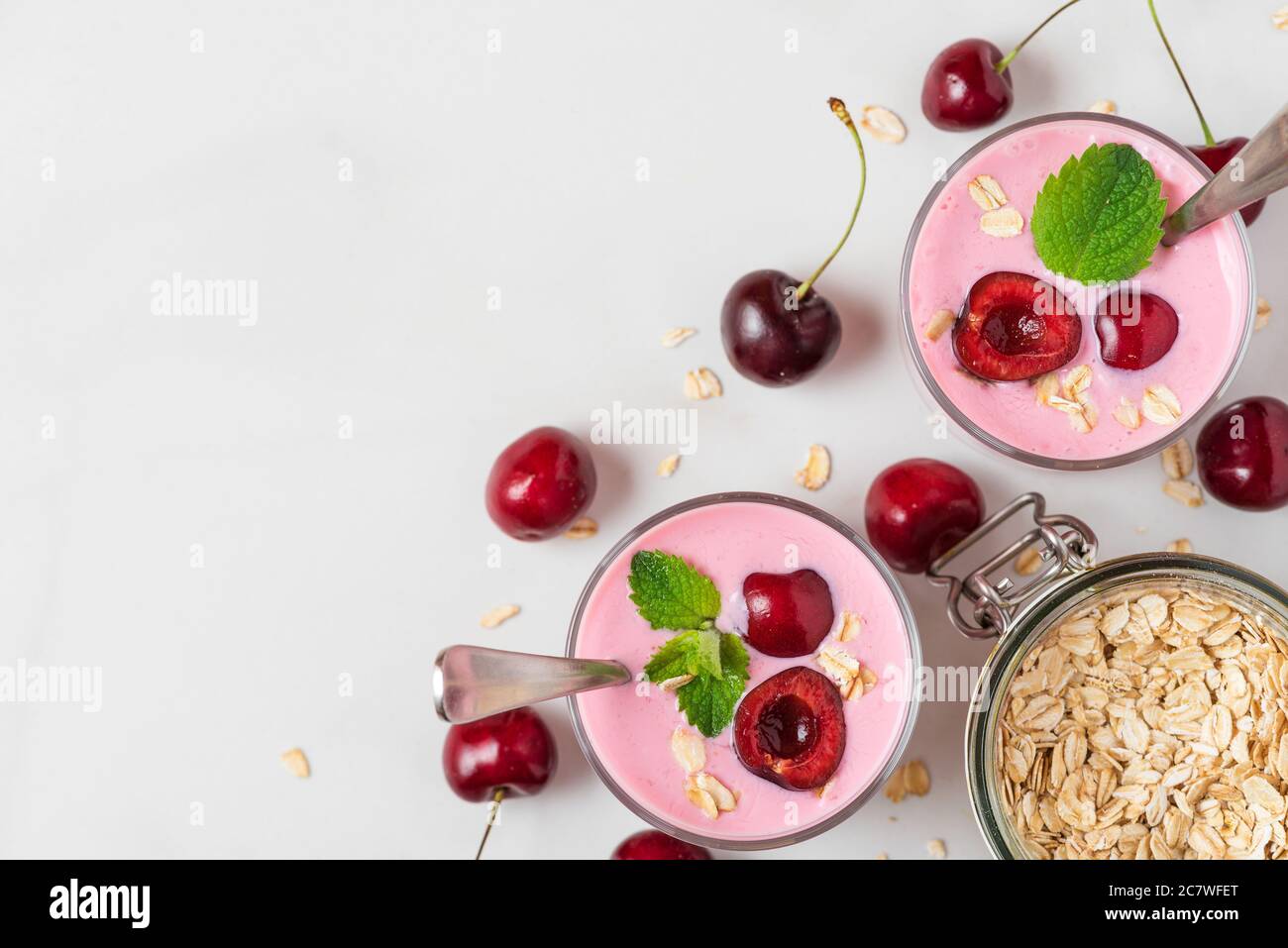 Yogurt di ciliegia con frutti di bosco freschi, avena e menta in bicchieri con cucchiai su sfondo bianco. Colazione salutare con vista dall'alto e spazio per la copia Foto Stock