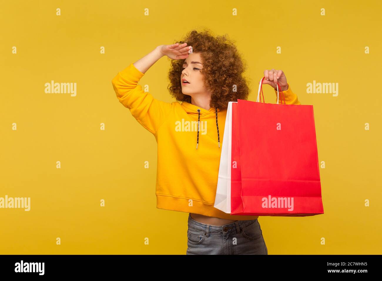 Ritratto di curly-capelli ragazza hipster curly-haired in felpa con cappuccio che tiene borse di shopping e guardando lontano, guardando la distanza, eyeing sconti in negozio. indo Foto Stock