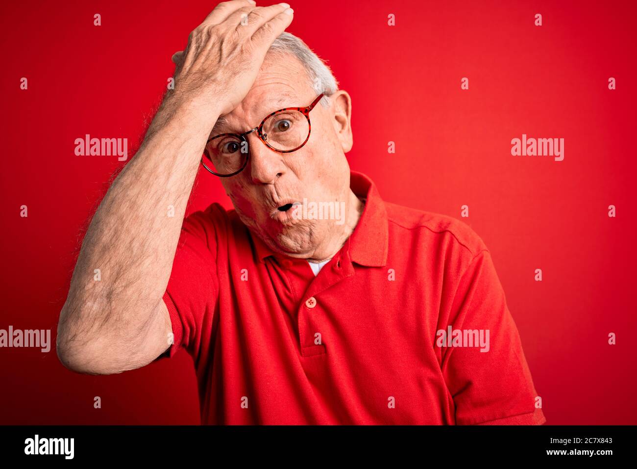 Uomo anziano con capelli grigi che indossa occhiali e t-shirt casual su sfondo rosso sorpreso con mano sulla testa per errore, ricorda l'errore. Dimenticato, Mem cattivo Foto Stock