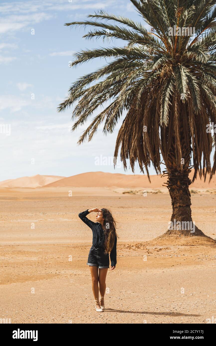 Donna che cammina nel bellissimo deserto con dune di sabbia e una sola palma. Viaggi in Marocco, Sahara, Merzouga. Libertà e concetto di viaggio. Foto Stock