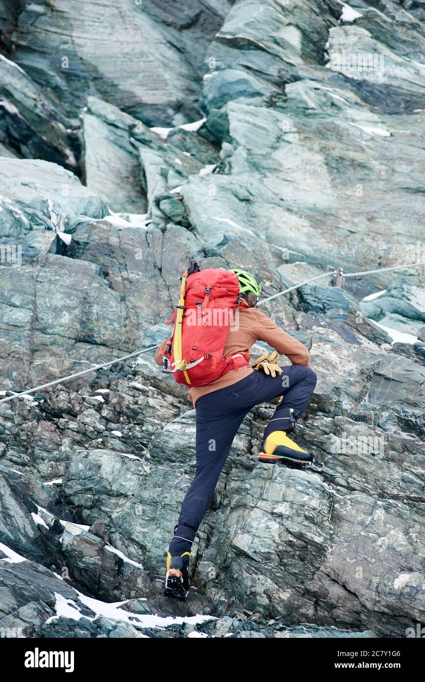 Vista posteriore dell'alpinista maschile con zaino usando la corda mentre si sale cresta alpina. Scalatore salita scogliera di montagna, cercando di raggiungere la cima. Concetto di alpinismo, alpinismo e alpinismo. Foto Stock
