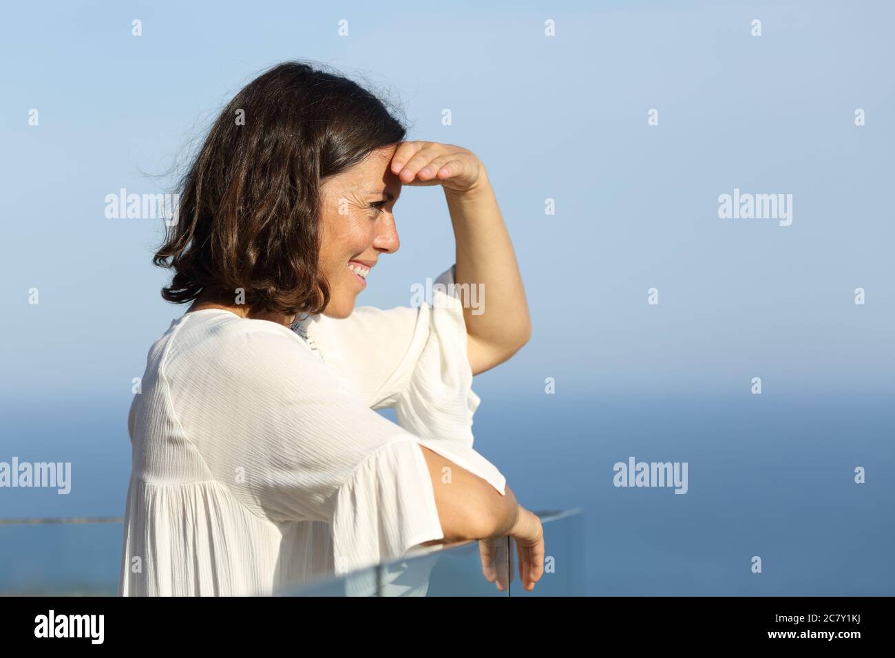 Vista laterale ritratto di una donna adulta felice che guarda le viste protegge dal sole in piedi in un balcone sulla spiaggia Foto Stock