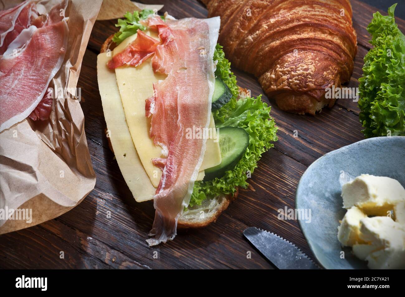 panino di croissant con fette di carne di jamon, foglie di lattuga verde, cetrioli freschi, formaggio, burro e coltello, su tavola rustica in legno, piatto Foto Stock
