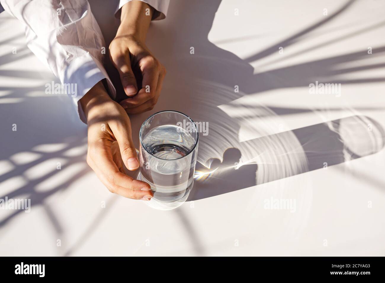 Le mani della donna e un bicchiere d'acqua pulita sul tavolo bianco in luce naturale con ombre vegetali. Foto Stock