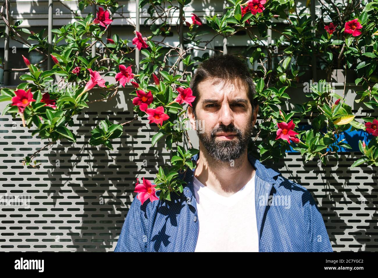 Uomo sopportato in abbigliamento casual poggiato su una parete fiorita mentre osserva la macchina fotografica Foto Stock