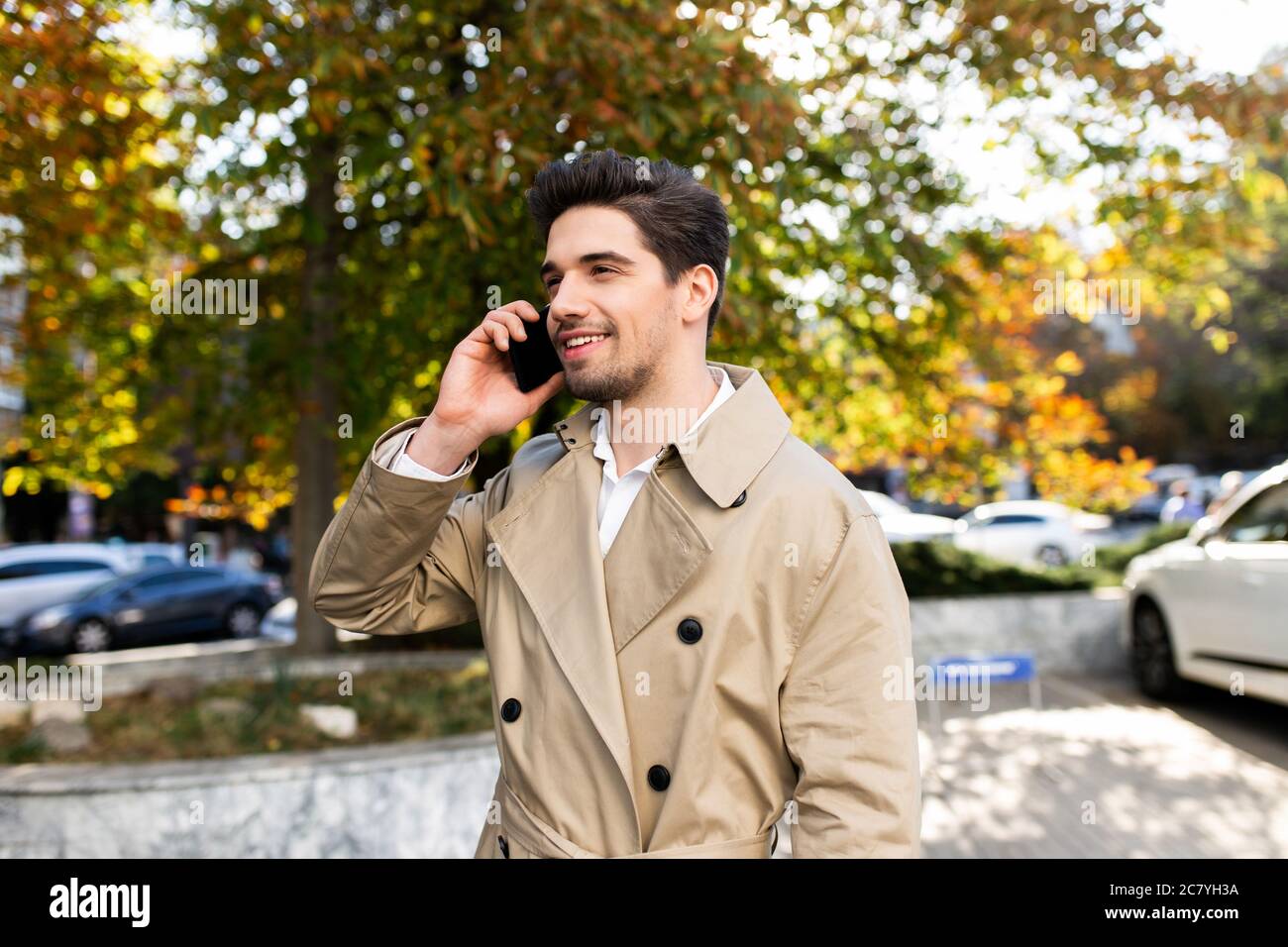 Giovane uomo attraente in trench cappotto felicemente parlando al cellulare all'aperto Foto Stock