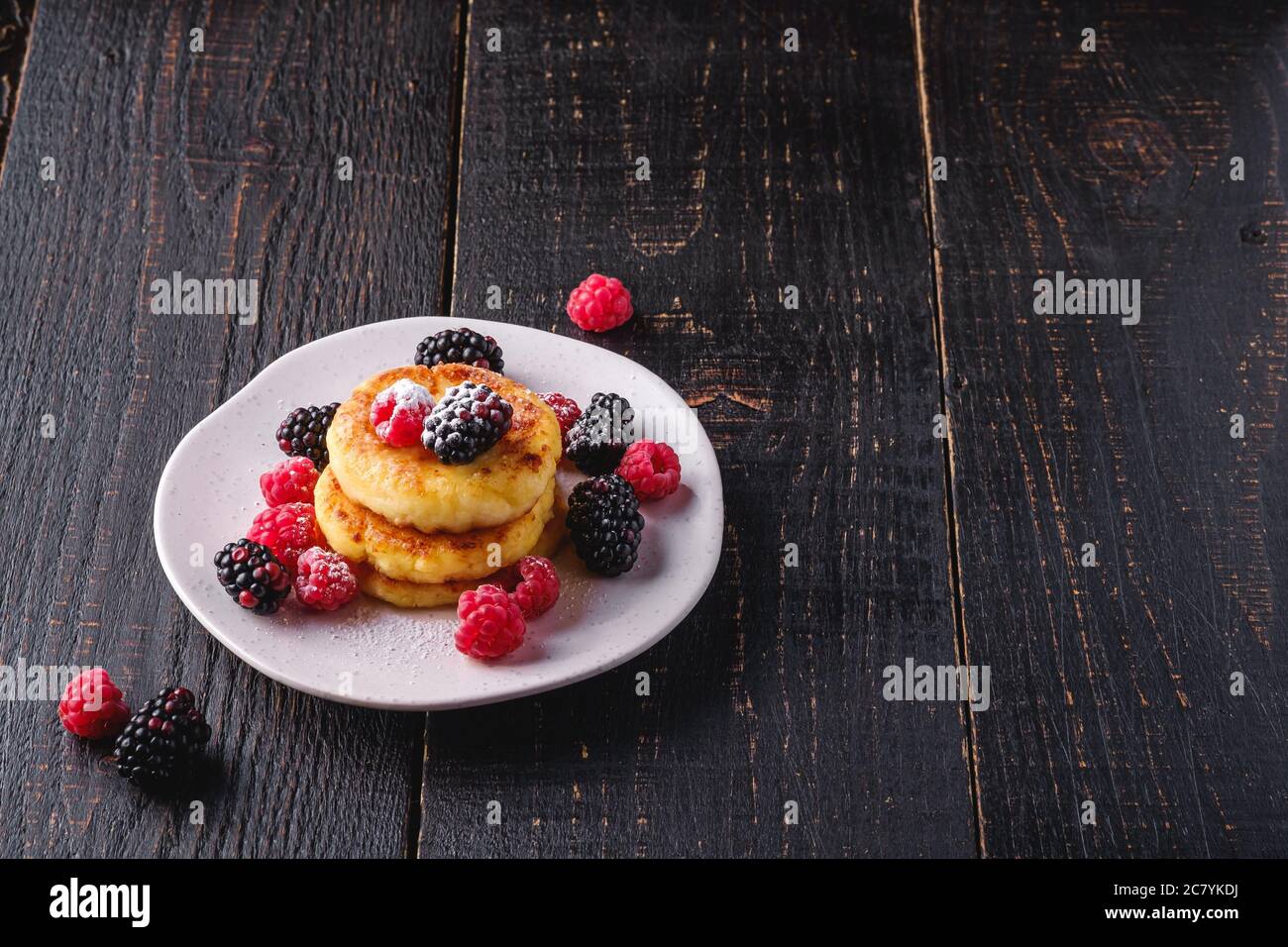 Frittelle di formaggio e zucchero in polvere, frittelle di cagliata dessert con lampone e frutti di bosco in piatto su sfondo nero scuro di legno Foto Stock