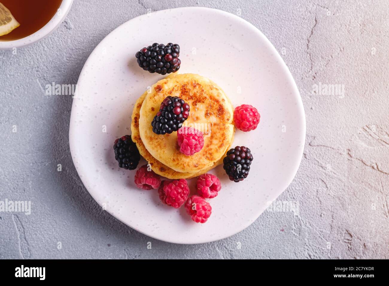 Frittelle di formaggio cottage, frittelle di cagliata dessert con lampone e frutti di bosco in piatto vicino alla tazza di tè caldo con fetta di limone Foto Stock